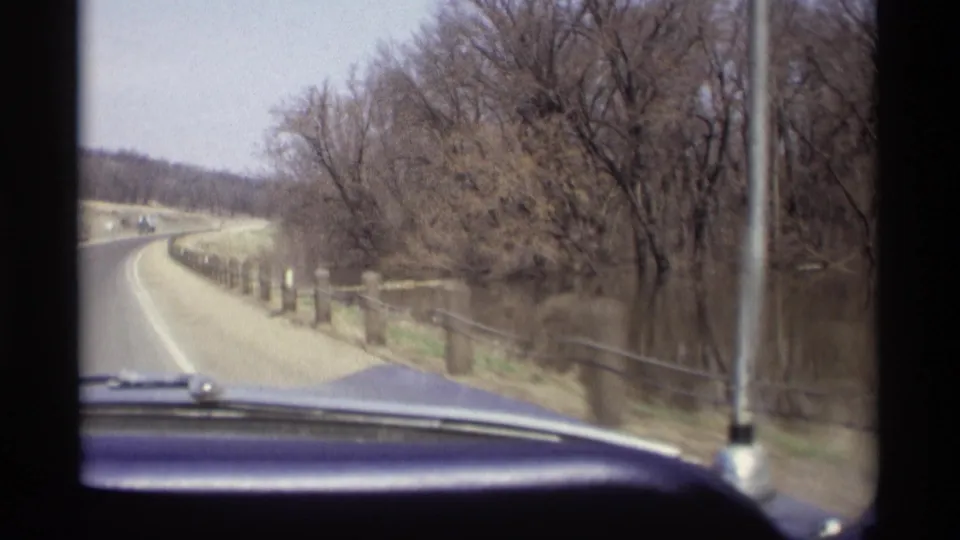 1969 Savage, Minnesota driving frame from inside a classic car, with a curving roadside, water, bare trees, and snow remnants visible.