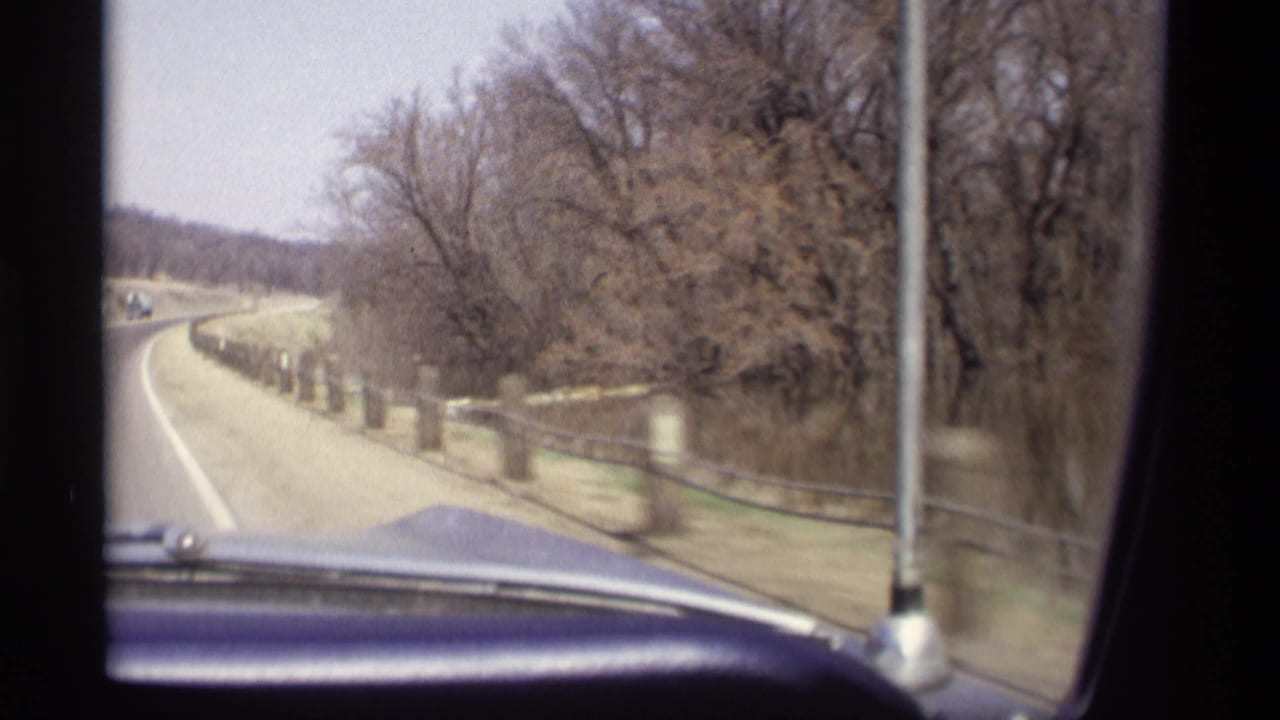 View from inside a car on a rural road bordered by posts, fences, and bare trees.