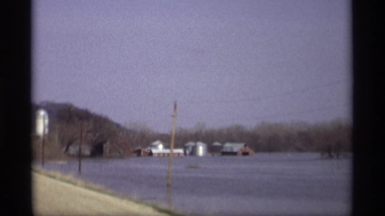 Flooded field with farm buildings, silos, bare trees, and a roadside utility pole.