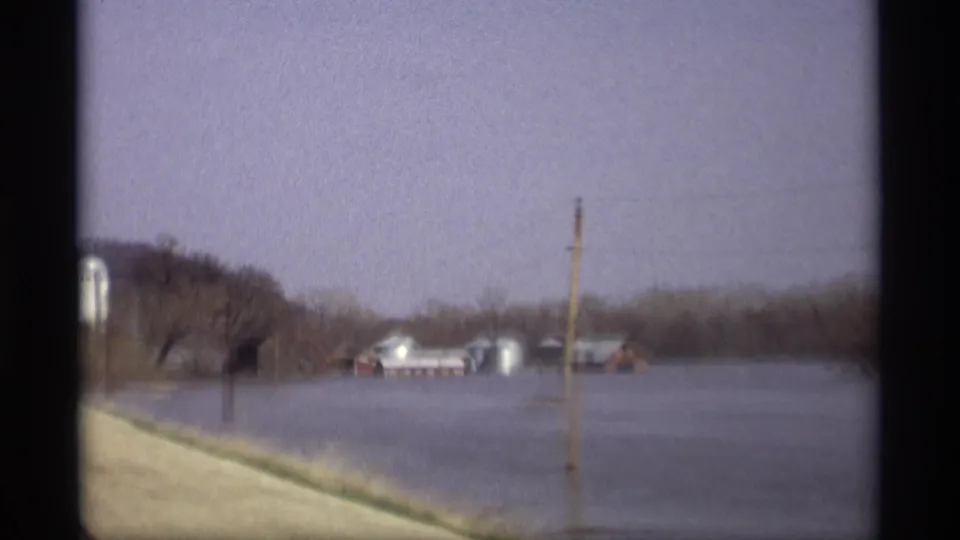 1969 Savage, Minnesota home-movie frame showing a roadside pass by flooded farmland with a pole, barns, silos, and wooded hills.