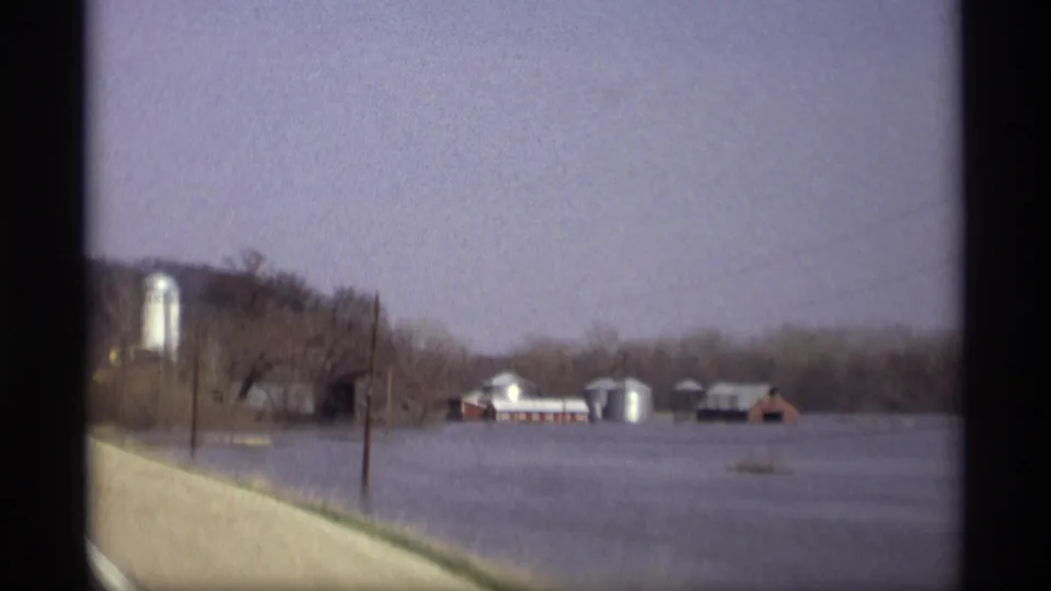 1969 Savage, Minnesota view of a flooded farmstead from a moving vehicle, with silos, barns, poles, and winter-bare trees visible.