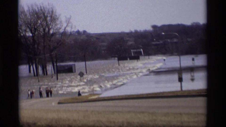 1969 Minnesota home-movie view of floodwater near a roadside, bare trees, and distant people standing at the water's edge.