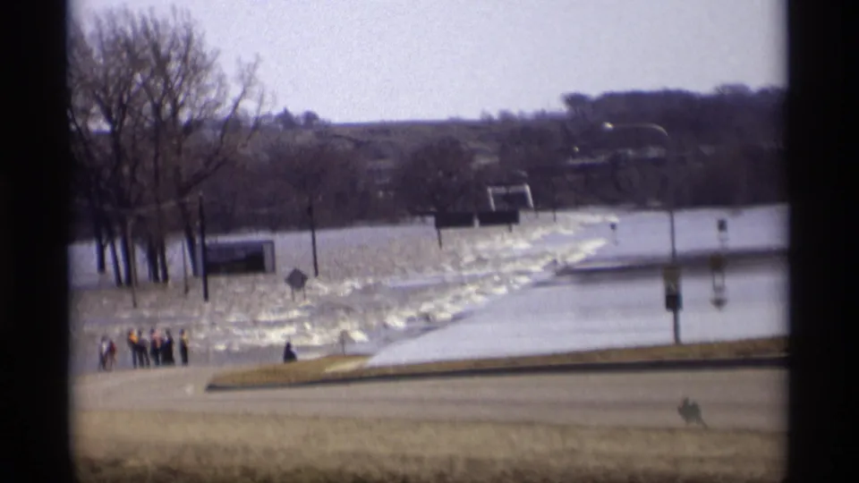 1969 Minnesota 8mm frame showing floodwater across a low rural area, with bare trees, roadside markers, and small distant figures.