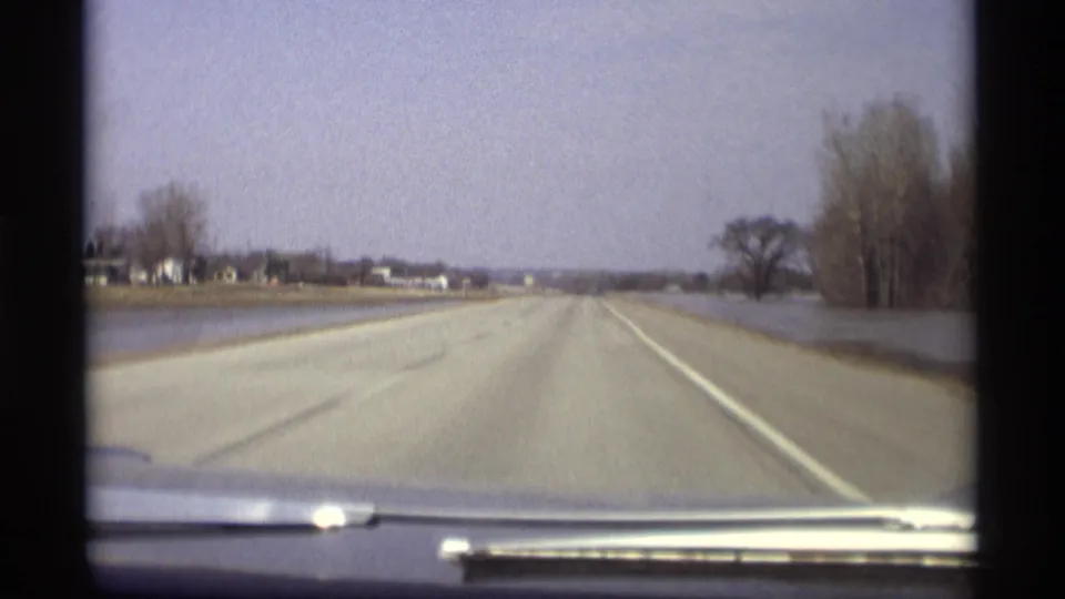 1969 Savage, Minnesota 8mm view over a car dashboard toward an empty rural highway bordered by floodwater.