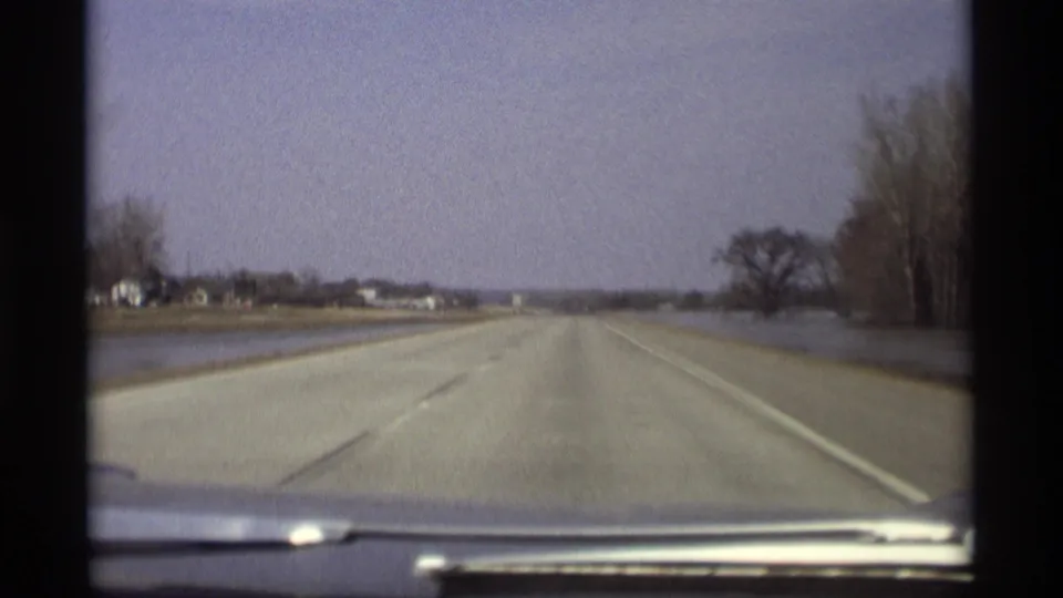 Dashboard view along a rural Savage, Minnesota highway with floodwater beside the road and distant buildings on the horizon.