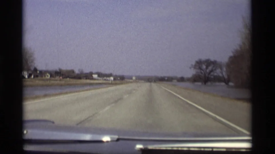 1969 home-movie frame from inside a car on a rural Minnesota highway with water pooled beside the pavement.