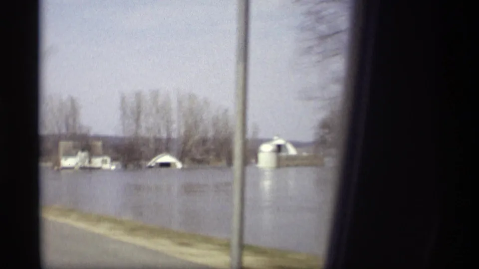 Rural Savage, Minnesota flood scene with water around small buildings, bare trees, and a roadside signpost seen from a moving car.