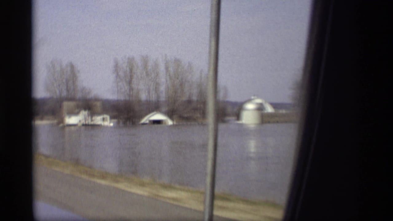 Floodwater surrounds low farm buildings and bare trees beside a road.