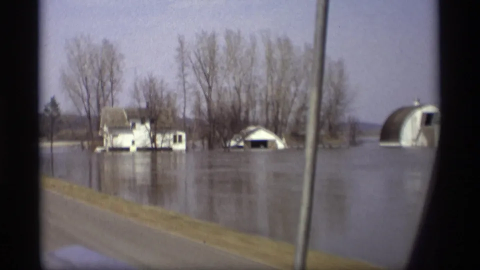 1969 Savage, Minnesota home-movie still showing floodwater around a house, shed-like structure, and arched building near bare trees.