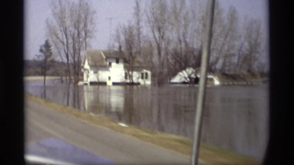 1969 Savage, Minnesota frame showing a white house, collapsed or low outbuilding, bare trees, and reflective floodwater beside the road.