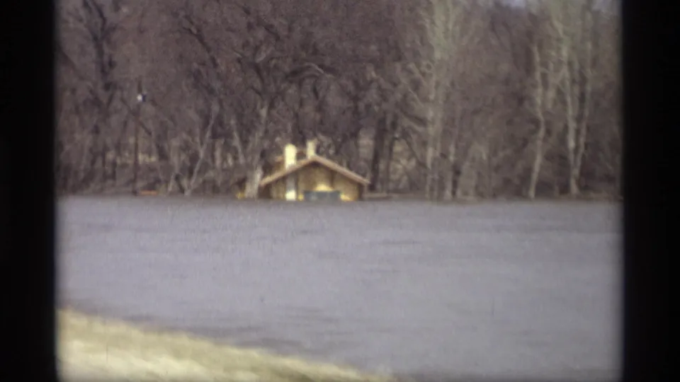1969 Savage, Minnesota home-movie frame showing murky floodwater in front of a small house or cabin near a wooded shoreline.