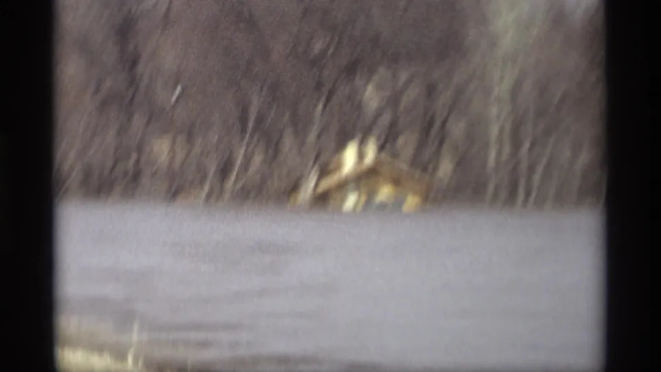 Motion-blurred 1969 Savage, Minnesota 8mm frame with floodwater, a small building, and bare trees seen from a moving roadside viewpoint.