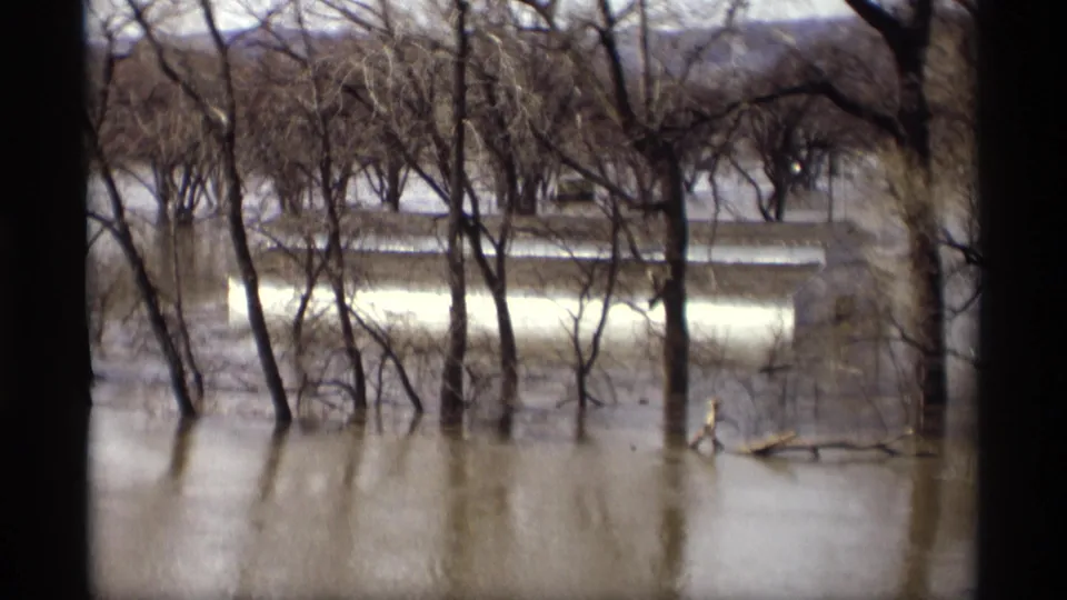 1969 Savage, Minnesota 8mm frame showing muddy floodwater around bare trees, with a pale house or structure partly visible in the background.