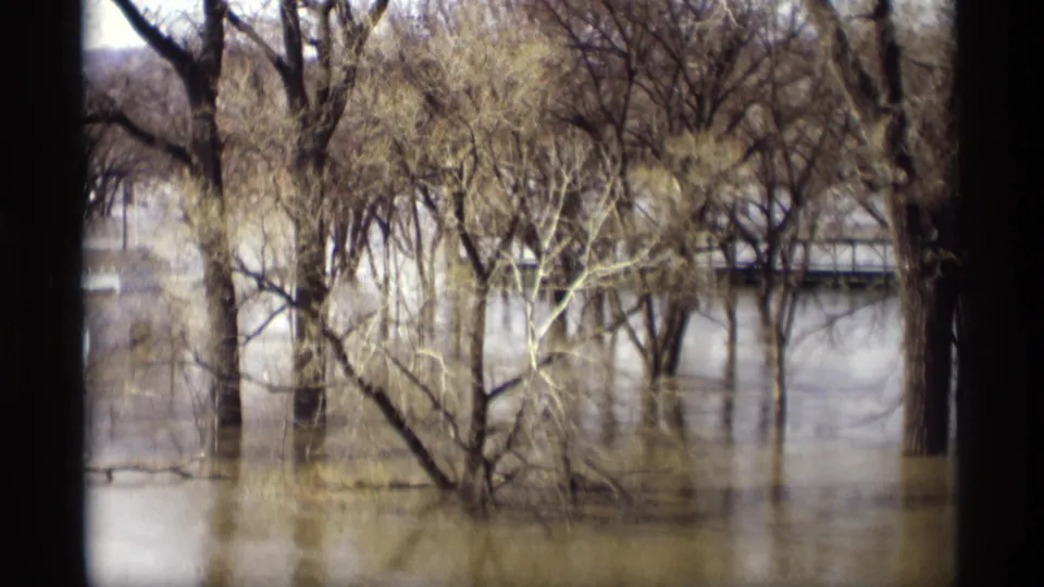 1969 Savage, Minnesota 8mm frame showing a flooded stand of bare trees, muddy water, and a low bridge or road edge in the background.