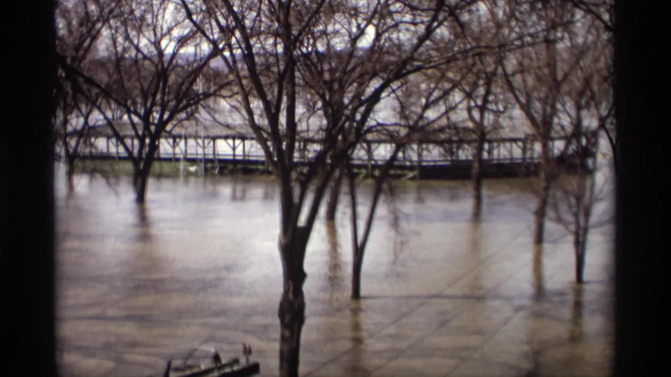 Vintage 8mm view of muddy floodwater around bare trees, with a partially submerged pedestrian bridge in the background.