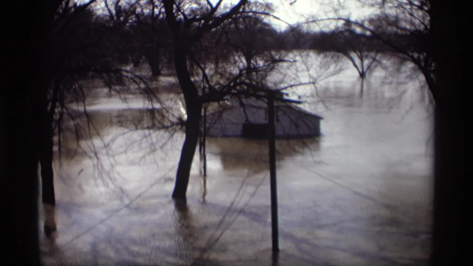 Floodwater nearly surrounds a small building in Savage, Minnesota, with bare trees and a dark foreground frame visible.