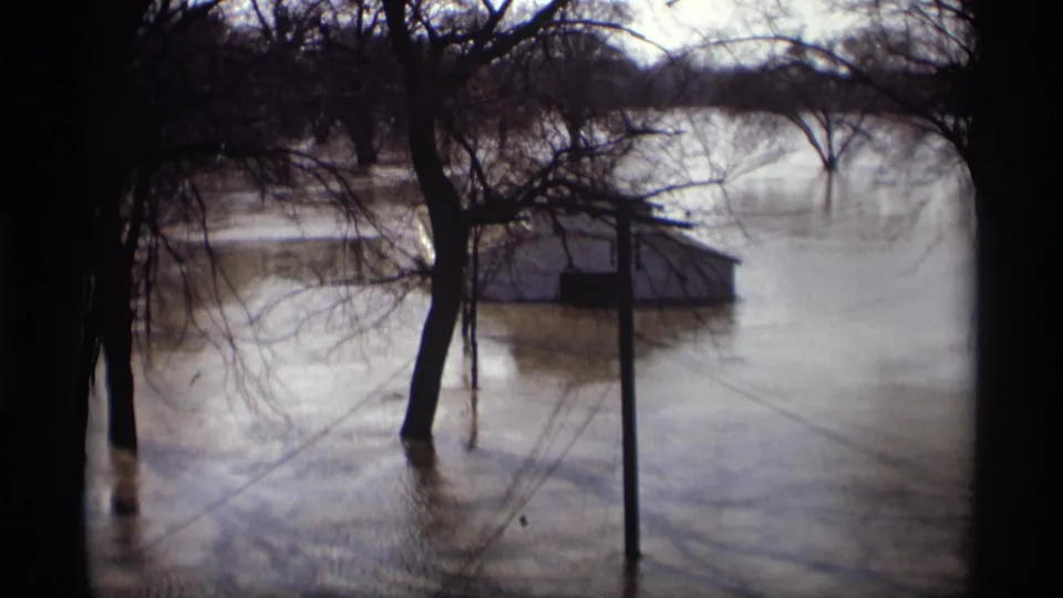 1969 Savage, Minnesota 8mm frame showing murky floodwater around leafless trees, a roadside post, and a low structure in the water.