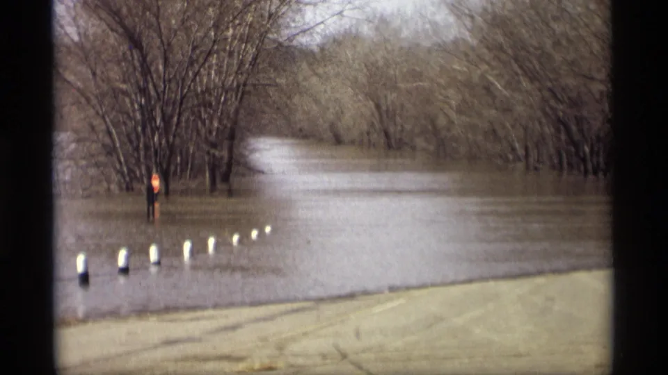 1969 Savage, Minnesota frame of floodwater crossing a roadside area, with white posts, bare trees, and a red sign partly surrounded by water.