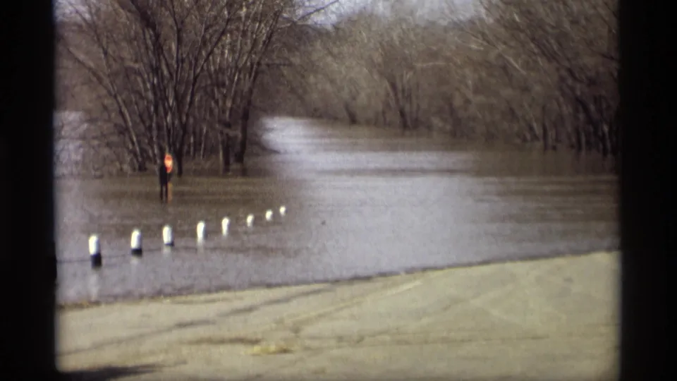 1969 Savage, Minnesota home-movie frame showing a flooded roadside lined with white posts, leafless trees, and a red sign in the water.