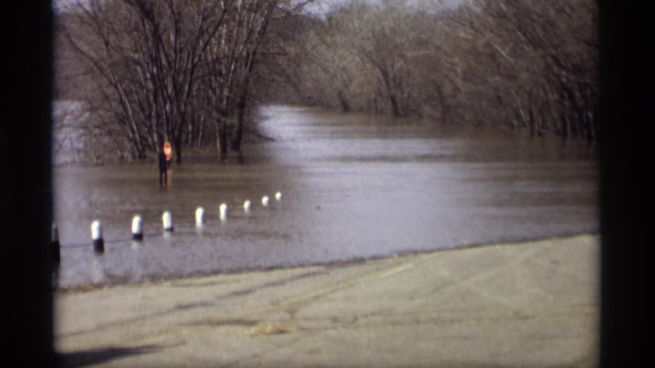 1969 Savage, Minnesota flood footage showing a submerged roadside, white bollards, bare trees, and a stop sign standing in high water.