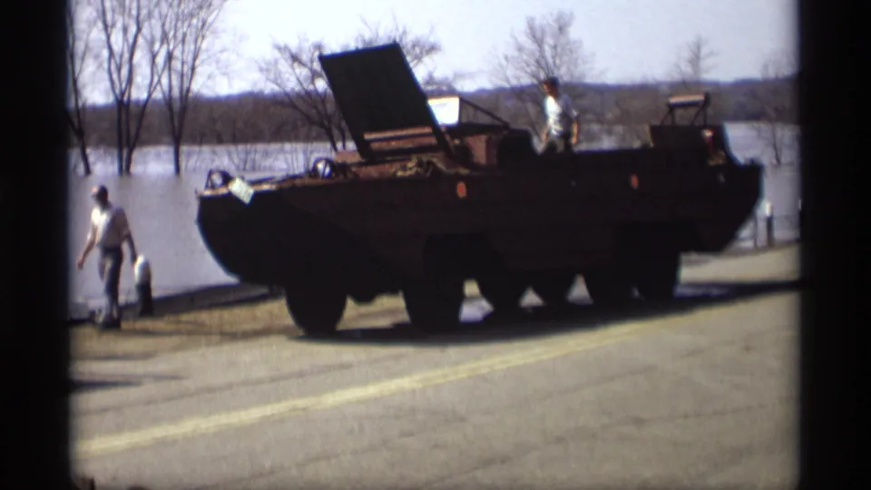 Men assess flood conditions near a military-style amphibious vehicle on a road in 1969 Savage, Minnesota home-movie footage.