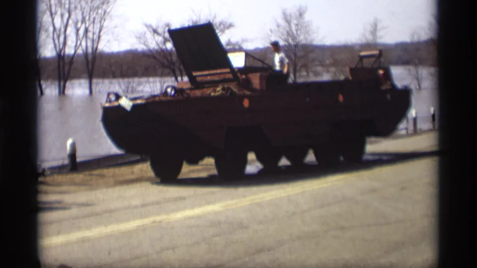 A military-style amphibious vehicle with an open hatch is parked on a flooded roadside in Savage, Minnesota in 1969 footage.