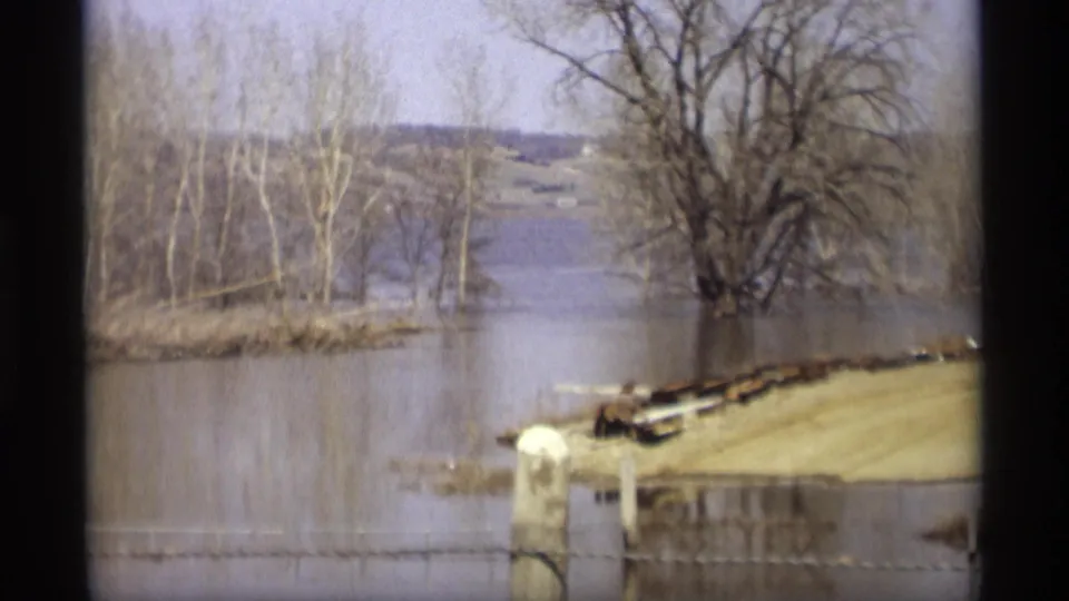 Savage, Minnesota floodwater surrounds a road or landing area, with bare trees, white posts, and a distant water channel in view.