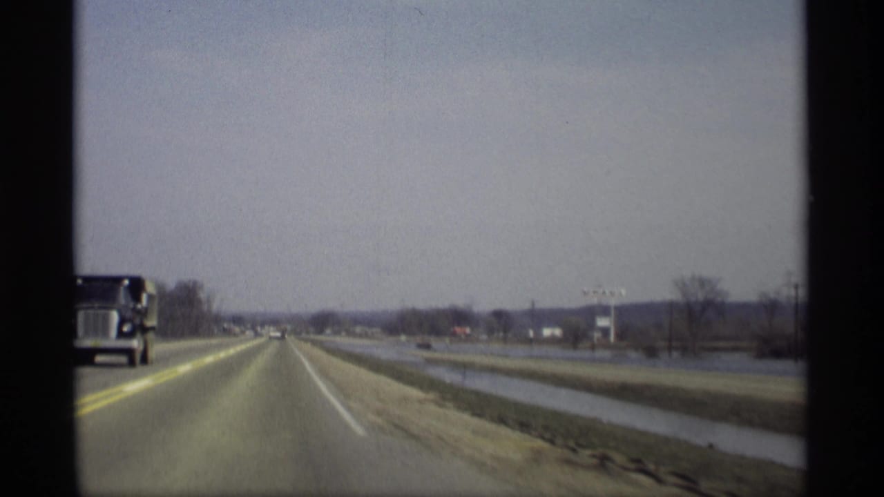 Two-lane rural highway seen from a moving vehicle with a large truck at left and water beside the road.