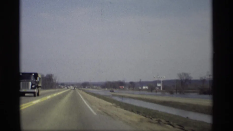 1969 8mm view from a moving vehicle on a rural highway near Savage, Minnesota, with a truck, cars ahead, wet fields, and bare trees.