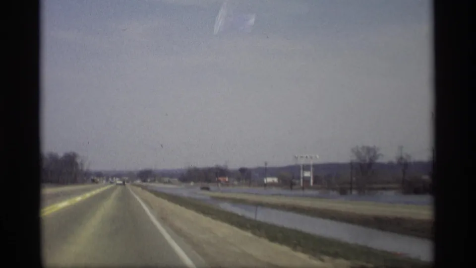 Forward-looking 1969 8mm road view near Savage, Minnesota, showing cars ahead, roadside water, open fields, utility poles, and distant structures.