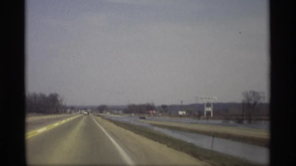 1969 home-movie frame from a vehicle on a rural Savage, Minnesota highway, with cars ahead, water by the shoulder, bare trees, and low buildings.