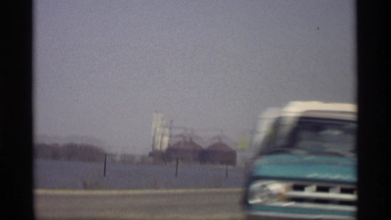 1969 Savage, Minnesota road view from a car, showing open water, distant industrial buildings, and a blurred vehicle passing in the foreground.