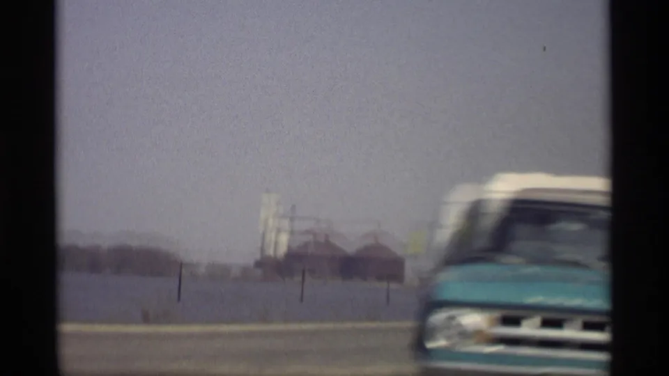 1969 Savage, Minnesota road view from a car, showing open water, distant industrial buildings, and a blurred vehicle passing in the foreground.