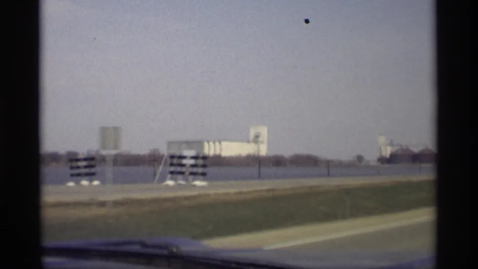 1969 Savage, Minnesota highway view from inside a moving car, with water, roadside signs, distant silos, and industrial buildings visible.
