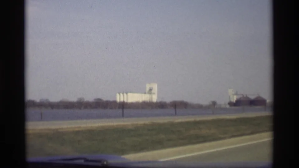 1969 Savage, Minnesota roadside view with a car hood edge, grassy shoulder, open water, distant silos, and low industrial structures.