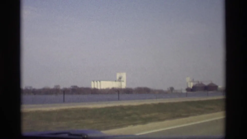 1969 Savage, Minnesota highway-side view showing grass, open water, distant grain elevators or silos, and industrial buildings.