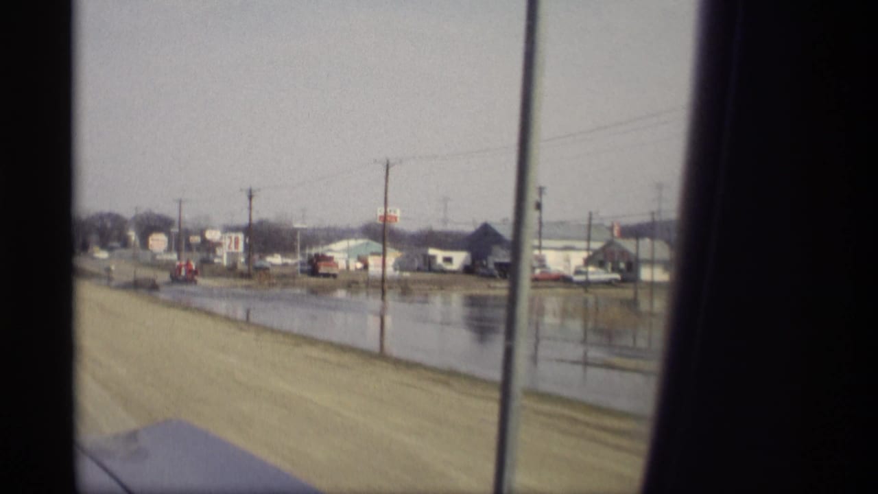 1969 Savage, Minnesota driving view past roadside flooding, commercial buildings, signs, utility poles, and a distant car.
