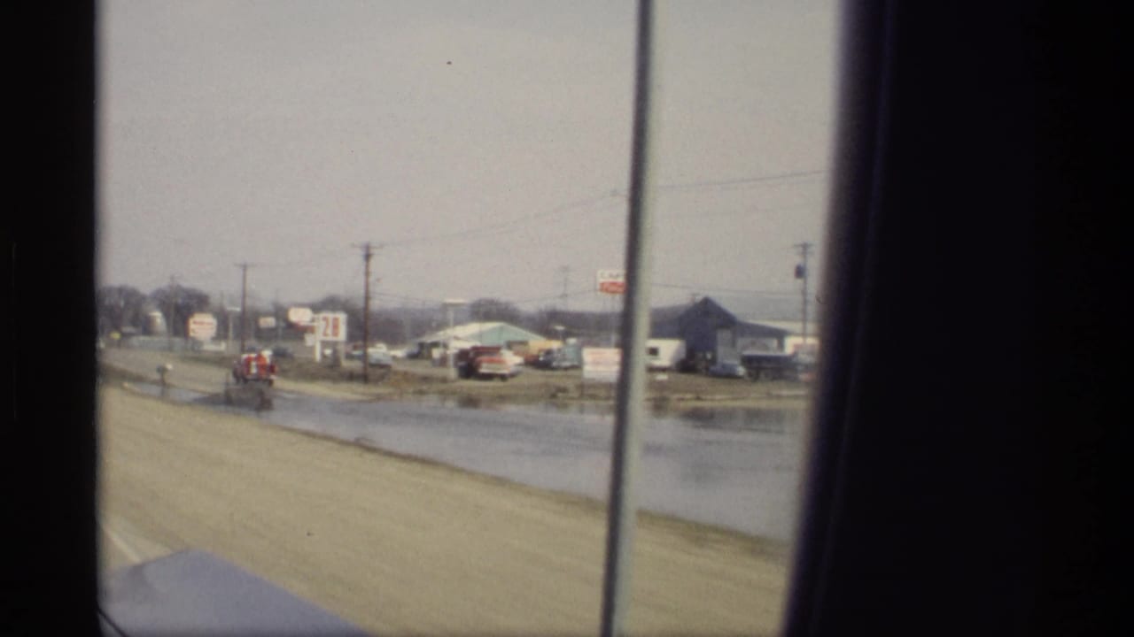 Moving car view of water beside the road, low buildings, signs, and traffic in the distance.