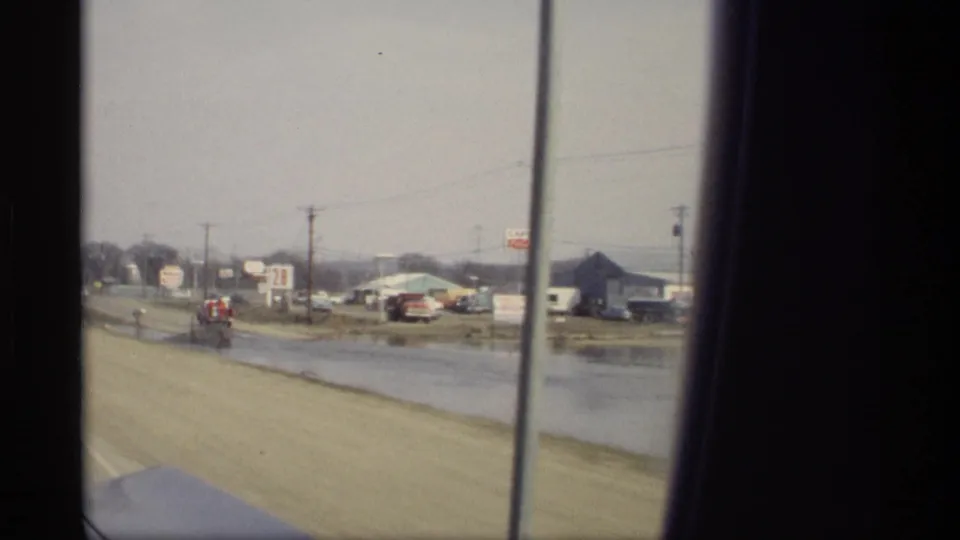 1969 Savage, Minnesota passenger-window view showing flooded roadside, low commercial structures, signs, and distant vehicles.