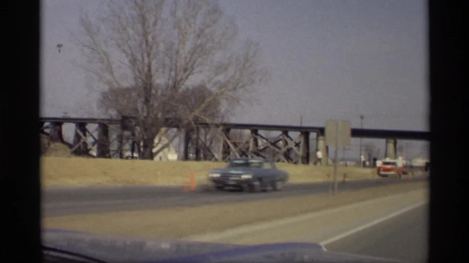 1969 Savage, Minnesota road view with a blurred blue sedan, bare trees, roadside cones, and a large wooden trestle bridge.