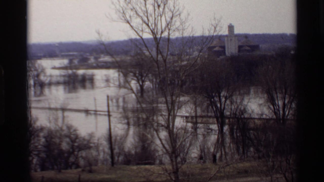 1969 Savage, Minnesota flood scene with water covering low land, bare trees in front, and distant buildings above the waterline.