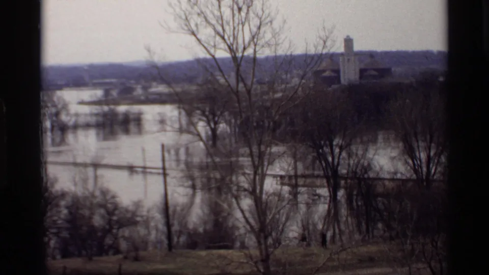1969 Savage, Minnesota flood scene with water covering low land, bare trees in front, and distant buildings above the waterline.