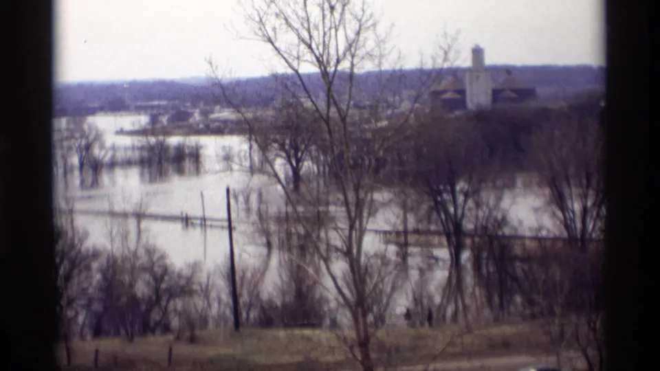 A handheld 8mm view of floodwater around trees and utility poles in Savage, Minnesota, with distant buildings on higher ground.