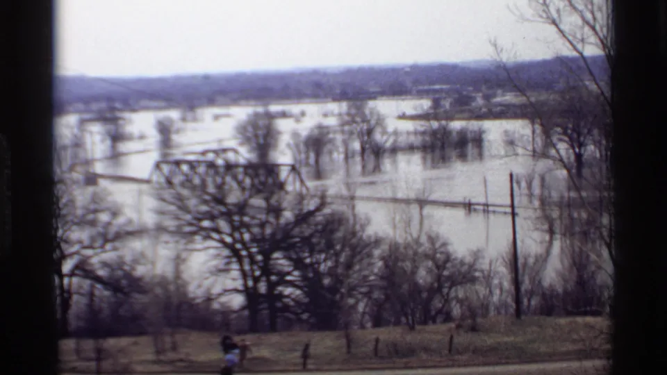 Savage, Minnesota floodwater covers land near a metal truss bridge, with bare trees and small distant people beside a road.