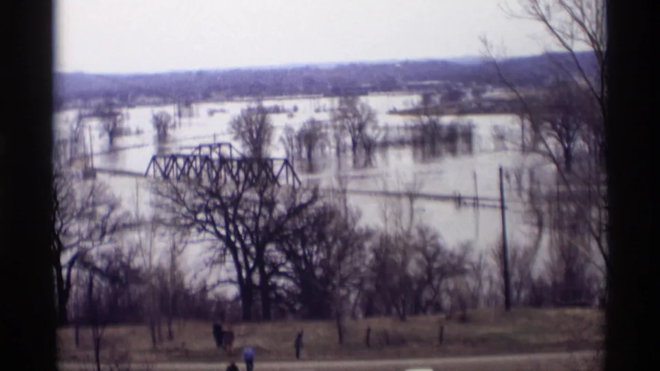 1969 8mm frame from Savage, Minnesota showing a truss bridge partly surrounded by floodwater, with trees and small people near the road.