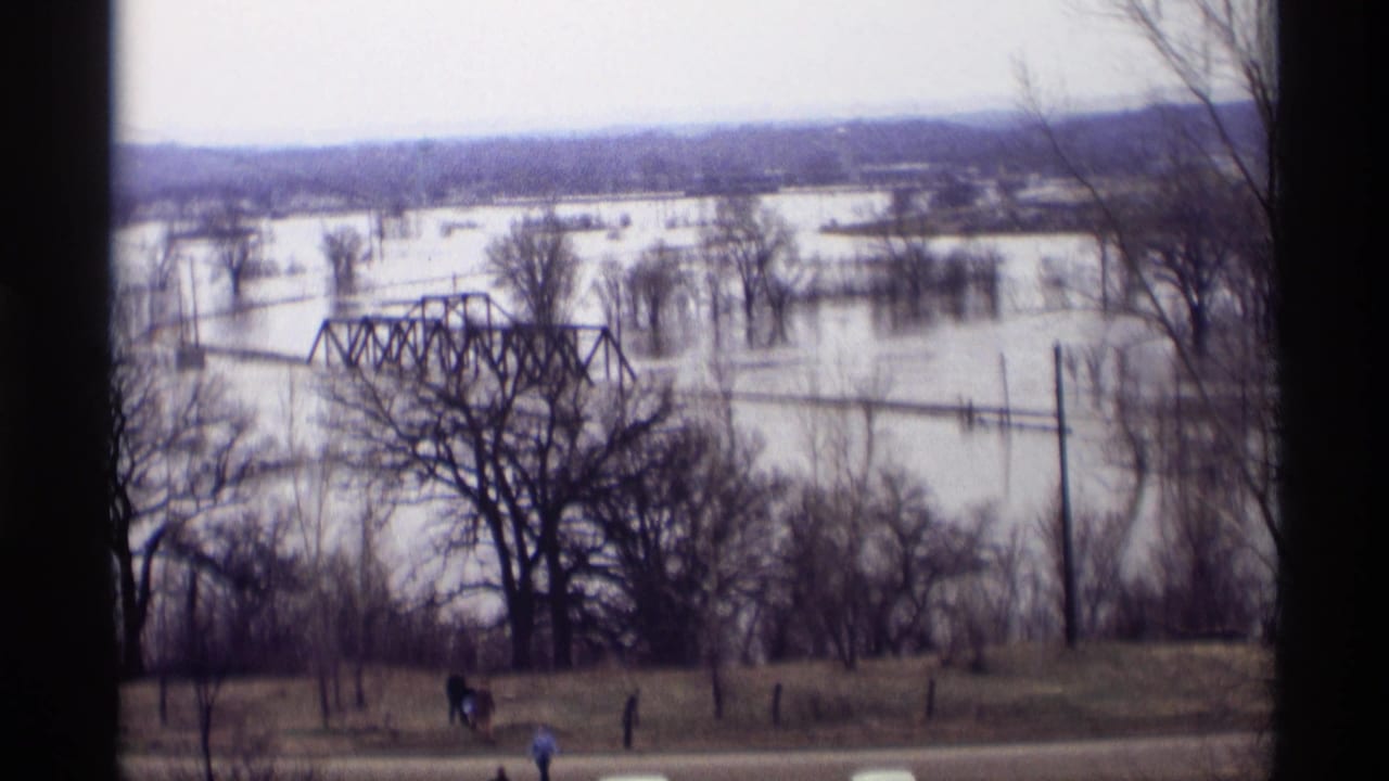 Flooded river valley with a steel truss bridge, bare trees, hillside observers, and road traffic below.