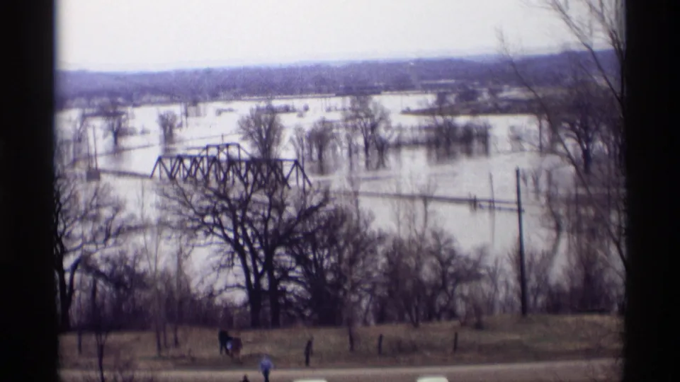 1969 Savage, Minnesota home-movie frame showing a flooded river valley, partly submerged truss bridge, bare trees, and people watching from a hillside.
