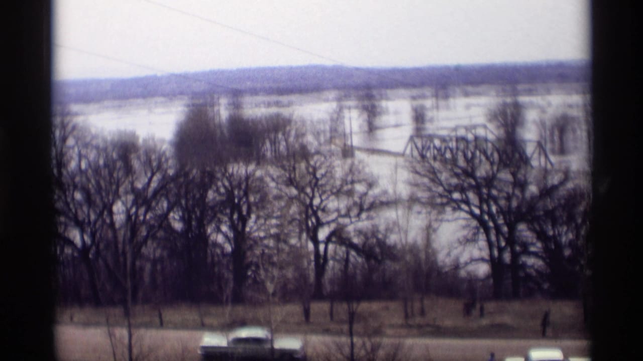Floodwater spreads around bare trees and a truss bridge, with a car moving along the road below.