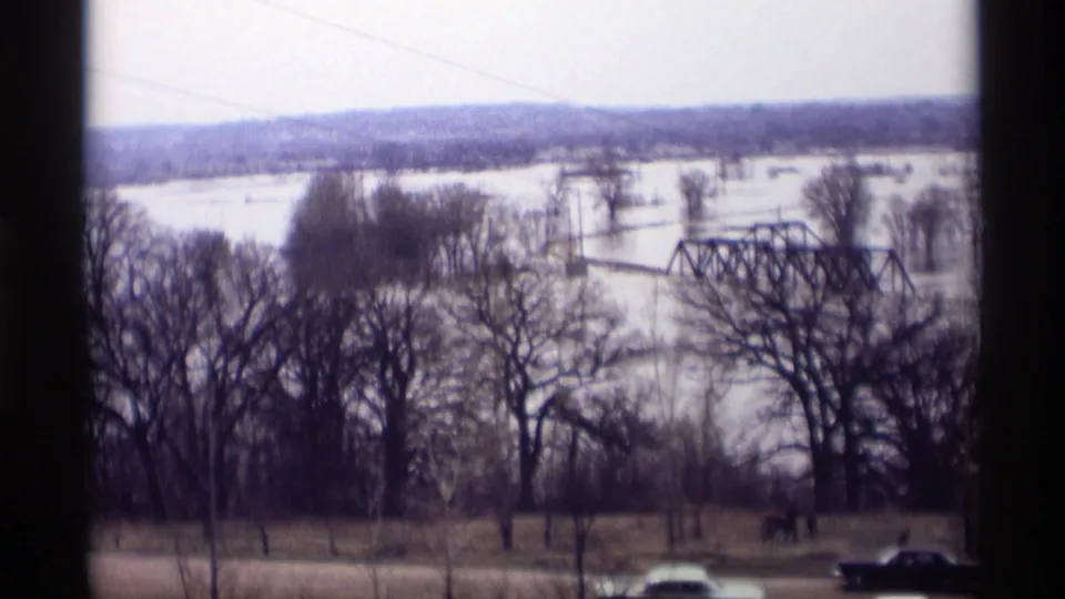 Soft 8mm frame from Savage, Minnesota in 1969, looking across a flooded winter river area with a truss bridge, bare trees, poles, and passing cars.