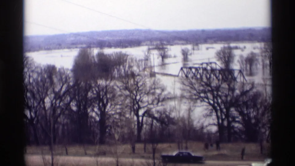 1969 Savage, Minnesota flood view showing water across the river flats, a steel truss bridge, winter trees, cars on the road, and distant observers.